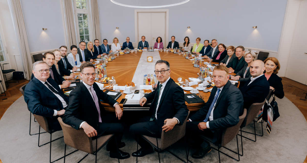 Die Landesregierung vor ihrer ersten Kabinettssitzung in der Villa Reitzenstein. Foto: Staatsministerium BW | Ilkay Karakurt 27 Mitglieder der Landesregierung sitzen um einen großen ovalen Tisch, im Vordergrund Cem Özdemir und Manuel Hagel.